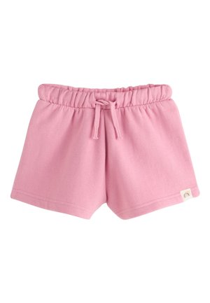 Junges Mädchen in rosa Sweatshirt, passenden Shorts, gestreiftem Hut und pinken Sneakern steht und schaut lächelnd nach oben mit den Händen in den Taschen.