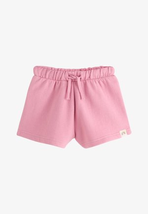 Junges Mädchen in rosa Sweatshirt, passenden Shorts, gestreiftem Hut und pinken Sneakern steht und schaut lächelnd nach oben mit den Händen in den Taschen.