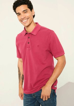 Fuchsia polo shirt with a collar, three buttons, and a small logo. Short sleeves and soft texture, paired with blue jeans.
