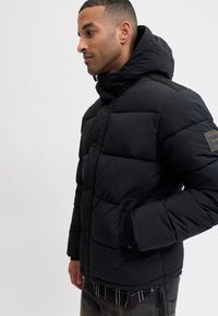 Chaqueta acolchada negra con cuello alto, botones a presión y bolsillos laterales. Presenta un tejido texturizado y un parche de la marca en la manga.