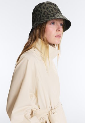 Vrouw met lang blond haar, die een beige mantel met ceintuur en een groene hoed met luipaardprint draagt, kijkt naar rechts met een neutrale uitdrukking.