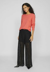 Koral strikkegenser med rund hals, kombinert med sorte blonder bukser med vide ben. Buksene har et blomstermønster og gjennomsiktig tekstur. Sorte spisse sko.