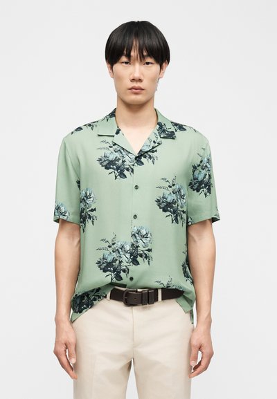 Νεαρός άνδρας που φοράει ανοιχτοπράσινο πουκάμισο με κοντά μανίκια και μαύρο-μπλε λουλουδάτο σχέδιο, μπεζ παντελόνι και σκούρο καφέ ζώνη.