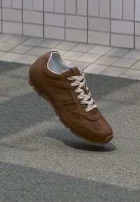 Brauner Sportschuh mit weißen Schnürsenkeln, der über einer strukturierten Asphaltoberfläche und einem gefliesten Wandhintergrund schwebt.