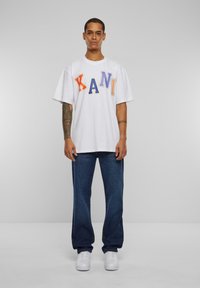 White oversized t-shirt with multicolored lettering spelling "KANI." Paired with dark blue jeans and white sneakers. Simple, casual design.