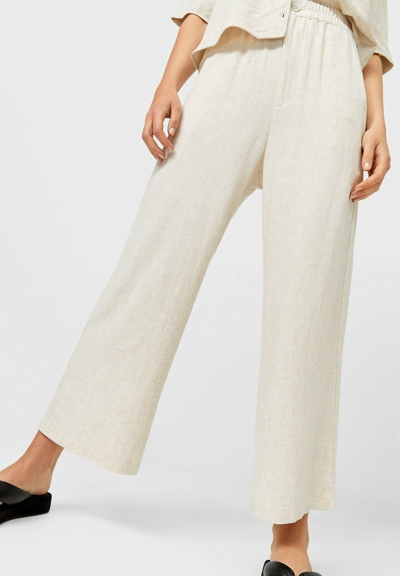 Persona che indossa pantaloni larghi e a vita alta in lino beige con una cintura elastica e scarpe nere slip-on su uno sfondo neutro.