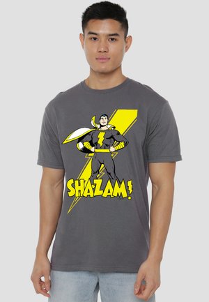 Man wearing gray T-shirt with yellow lightning bolt and superhero figure standing with hands on hips, text "SHAZAM!" below.