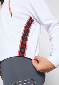 Valkoinen urheiluhuppari, jossa on lyhyempi malli, ja siinä on rohkea musta ja punainen FILA-logo raita sivussa ja naruakoristeet.