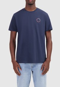 Donkerblauwe katoenen t-shirt met ronde hals, korte mouwen en een kleine cirkelvormige grafiek op de linkerborst in rood en wit.