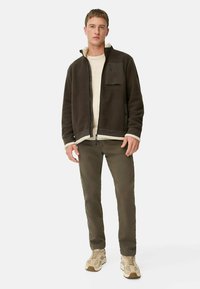 Brun fleecejakke med høj krave, lynlås lukning og en brystlomme. Sammen med olivengrønne bukser og beige sneakers.