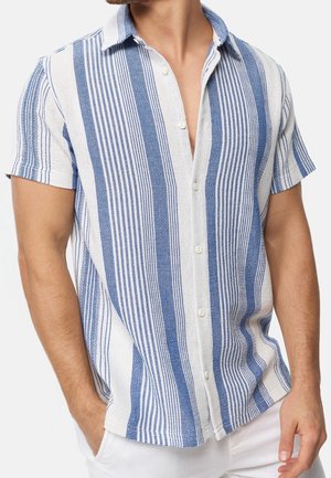 Man wearing short-sleeve button-up shirt with vertical blue and white stripes, and white pants with one hand in pocket.