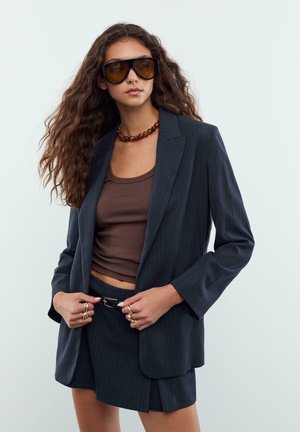 Žena s dlouhými kudrnatými vlasy, která nosí velké tmavé sluneční brýle, hnědé tílko, tmavý pruhovaný blazer, odpovídající sukni a několik prstenů.