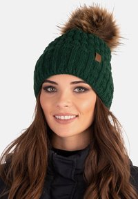Gorro de punto verde con un patrón texturizado y un pompón de piel marrón en la parte superior. Presenta una pequeña etiqueta de marca en el lateral.