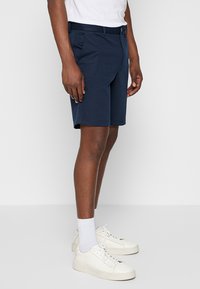 Navyblaue Herren-Shorts mit taillierter Passform, aus einem strukturierten Stoff, mit Seitentaschen und Knopfverschluss, getragen mit weißen Sneakers.