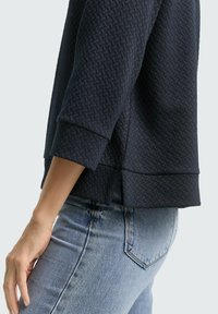 Navyblauwe, gestructureerde sweatshirt met een geometrisch patroon, voorzien van driekwart mouwen en een rechte zoom, gedragen over lichtblauwe jeans.
