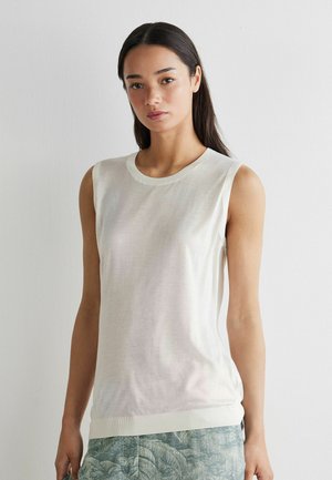 Vrouw met lang donker haar, gekleed in een mouwloos wit topje en groen met patroon broek, staand tegen een effen witte muur.