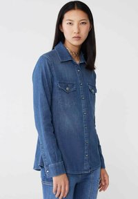 Camicia di denim blu scuro, con colletto a punta, due tasche sul petto con pattina, chiusura con bottoni e maniche lunghe con polsini a bottoni.
