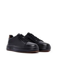 Un paio di sneakers in pelle nera con suole spesse e lacci neri, viste frontalmente da destra su uno sfondo bianco.