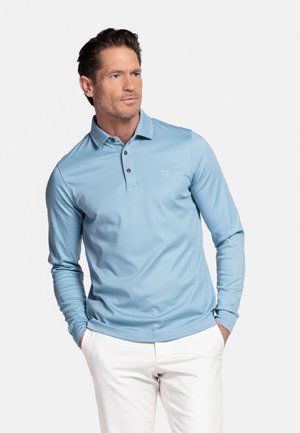 Man wearing light blue long-sleeve polo shirt and white pants, standing with hands in pockets, looking slightly to the side.