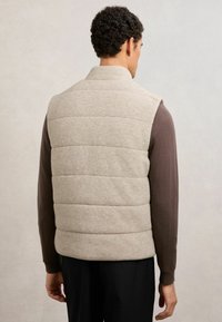 Mann von hinten gesehen, trägt eine beige gesteppte Weste über einem braunen Langarmshirt und schwarze Hose vor hellem, einfarbigem Hintergrund.