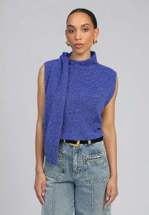 Femme portant un haut bleu en tricot sans manches avec détail d'écharpe attachée, associé à un jean en denim clair taille haute et des boucles d'oreilles créoles en or.