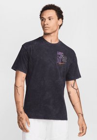 Man with tattoos wearing a black graphic t-shirt with number 72 and basketball net design, paired with white shorts, looking to the side.