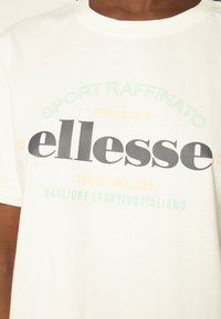 Weißes Baumwoll-T-Shirt mit rundem Halsausschnitt. Die Vorderseite zeigt mehrfarbigen Text: "Sport Raffinato", "ellesse" und italienische Phrasen in verschiedenen Schriftarten.