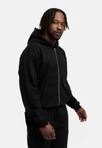 Schwarze Zip-Hoodie aus weichem Stoff, mit einer Fronttasche und Kordelzugkapuze, getragen mit passenden schwarzen Sweatpants.
