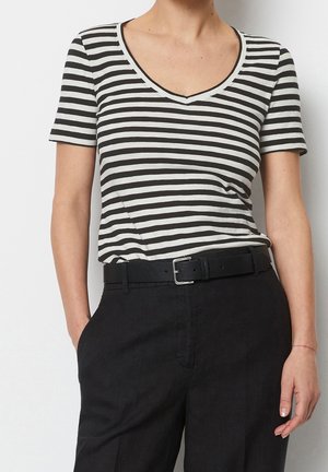 Person iført en sort og hvid vandret striped V-udskæring kortærmet skjorte stoppet ned i sorte bukser med sort bælte, hånd i lommen.