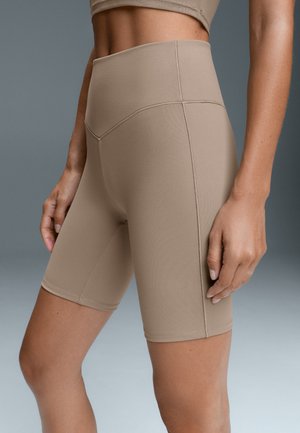 Femme portant un short de vélo beige taille haute et un haut assorti, montrée du ventre jusqu'à la mi-cuisse sur un fond gris.
