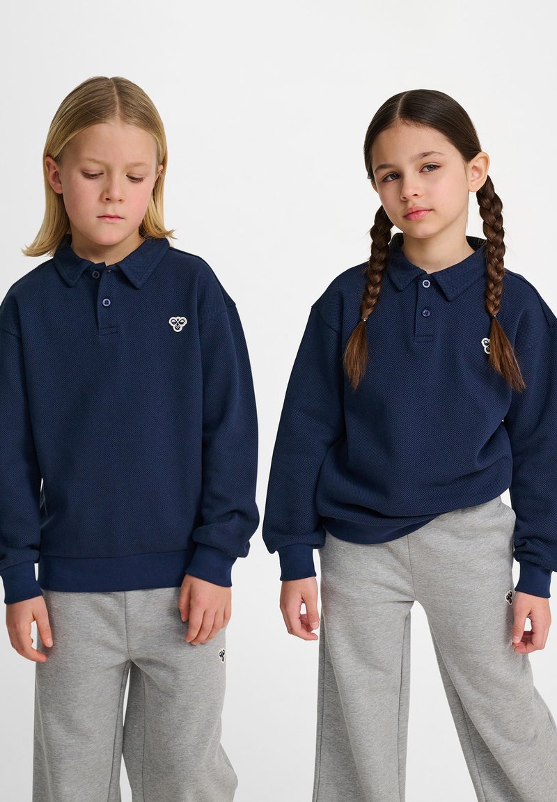 Två barn bär mörkblå sweatshirts med polo-kragar och grå joggingbyxor. Varje tröja har en logotyp på bröstet och en avslappnad passform.