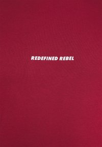 T-shirt en coton rouge avec un col rond, affichant en son centre un texte blanc audacieux « REDEFINED REBEL ». Texture lisse.