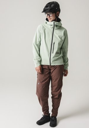 Person, die eine hellgrüne wasserdichte Jacke, braune Hose, schwarzen Fahrradhelm und Schutzbrille trägt und vor neutralem Hintergrund steht.