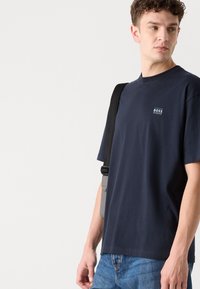 Tricou scurt cu mânecă, de culoare navy, fabricat din bumbac, cu guler rotund. Are un mic logo alb "BOSS" pe pieptul stâng. Este purtat cu blugi albaștri și o geantă crossbody.