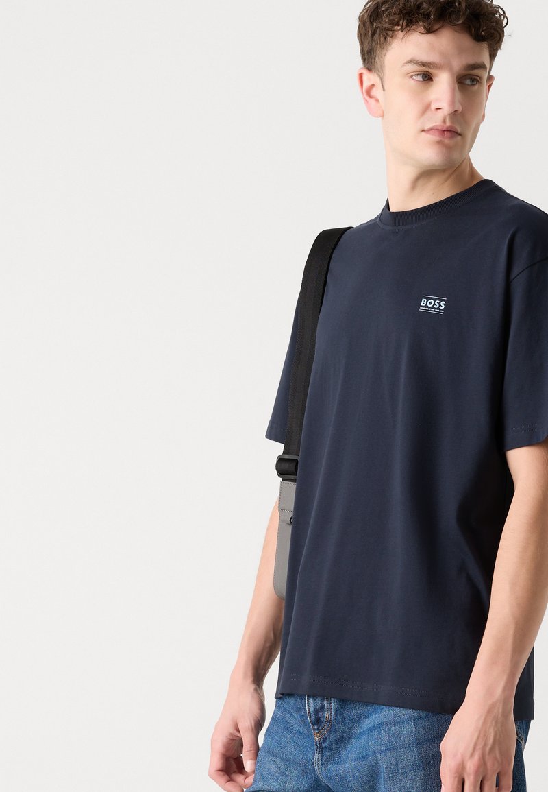 T-shirt navy a maniche corte in cotone con collo rotondo. Presenta un piccolo logo bianco "BOSS" sul lato sinistro del petto. Indossato con jeans blu e una borsa a tracolla.
