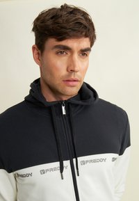 Felpa con zip bianca e nera con fascia a logo, dotata di cappuccio regolabile e texture morbida. Il corpo è nero, mentre le maniche sono bianche.