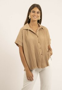 Chemise légère marron clair à manches courtes, avec une texture plissée verticale, un col et une coupe décontractée, associée à un pantalon blanc.