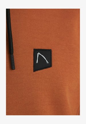 La tela del sudadera marrón presenta un parche cuadrado negro con un diseño gráfico en blanco. Incluye un cordón negro en el cuello.