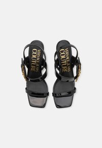 Sandali neri in vernice con punta quadrata, design a doppia cinghia e dettaglio con fibbia. Testo del logo dorato sulla soletta. Texture liscia.