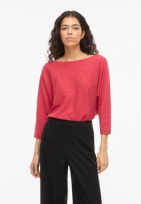 Vrouw met lang, krullend haar, gekleed in een rood shirt met lange mouwen en zwarte broek met hoge taille, staand tegen een witte achtergrond.