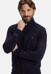 Homme avec une barbe et des cheveux bruns ondulés ajustant le col d'un pull zippé bleu marine sur un fond blanc uni.