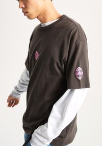 Camiseta de manga corta marrón oscuro con gráficos de calaveras rosas en el pecho y la manga, superpuesta a una camisa de manga larga gris claro.