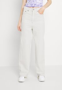 Jeans beiges de pierna ancha, confeccionados en denim, con una cintura alta, bolsillos frontales y una textura suave. Combinados con un top corto de tie-dye púrpura y zapatillas blancas.