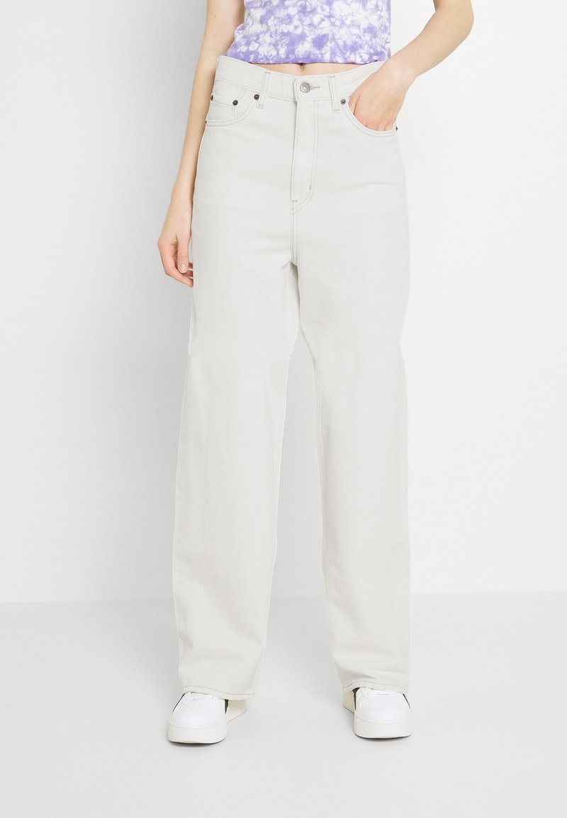 Jeans beiges de pierna ancha, confeccionados en denim, con una cintura alta, bolsillos frontales y una textura suave. Combinados con un top corto de tie-dye púrpura y zapatillas blancas.