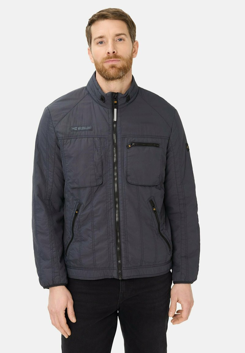 Man met kort haar en baard, gekleed in een donkergrijze doorgestikte jas met ritssluiting aan de voorkant en meerdere zakken, staand tegen een witte achtergrond.