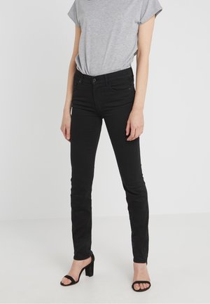 Person trägt schwarze Slim-Fit-Jeans, graues kurzärmeliges Oberteil und schwarze Sandalen mit Absatz und steht auf einem schlichten weißen Boden.