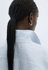 Cheveux tressés coiffés en une longue queue de cheval, portant une chemise col bleu clair en tissu texturé avec des coutures visibles. Boucles d'oreilles en argent.