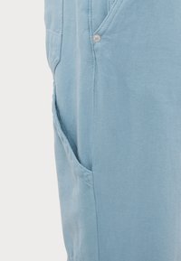Pantalon en denim bleu clair en gros plan montrant la poche avant avec rivet et détail de couture latérale sur fond uni.