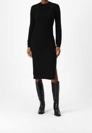 Mujer vestida con un vestido midi negro ajustado, de manga larga y con aberturas laterales, combinado con botas de cuero negras hasta la rodilla.