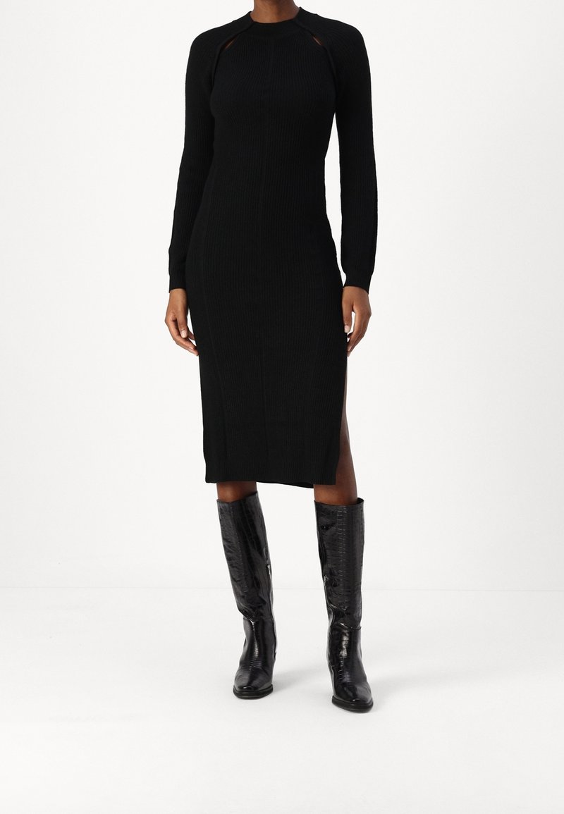 Femme portant une robe midi noire ajustée à manches longues avec des fentes sur les côtés, associée à des bottes en cuir noires montantes jusqu'aux genoux.